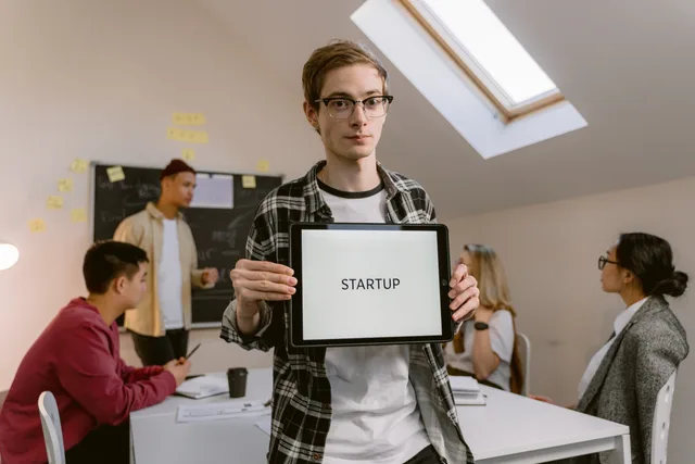 A young man in an office holding a tablet with 'STARTUP' during a meeting. Six startup funding outlooks for England in 2027