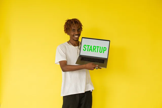 Smiling man holding a laptop with 'STARTUP' on screen against vibrant yellow background. Startup valuation methods in the UK: 5 approaches for first-time founders